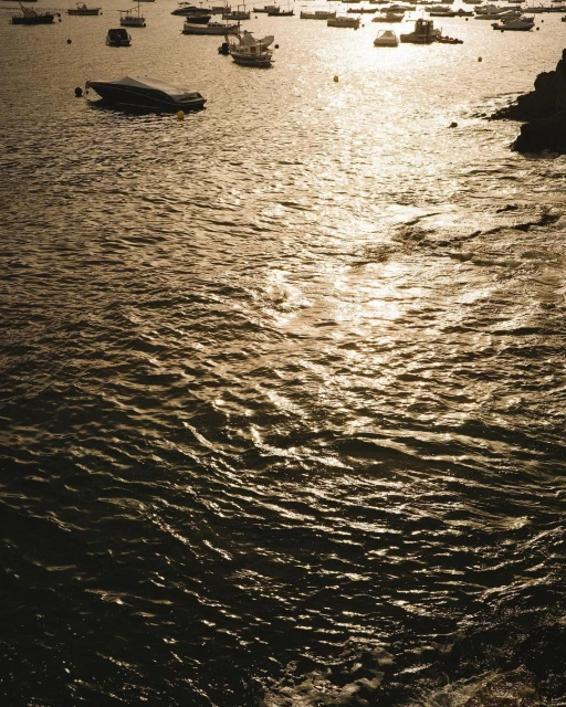 Golden sunlight glistens on the rippling ocean water near a rocky coastline, with several boats visible on the horizon at dusk.
