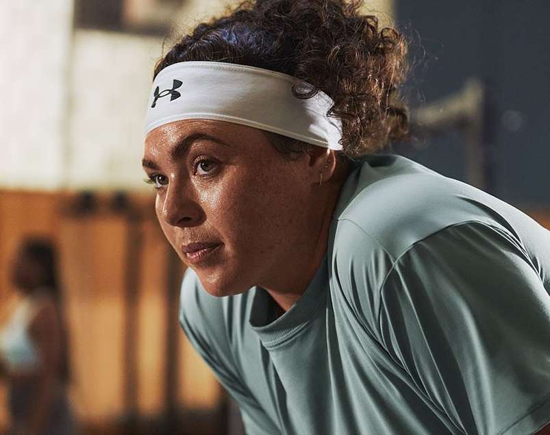 Adult female wearing a gray shirt and white headband with a logo, stands in a room with another person in the background.