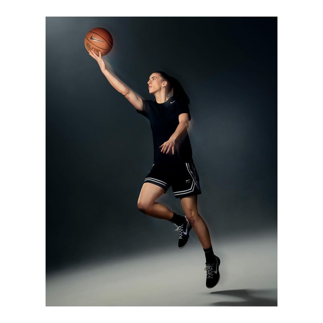 Athletic woman jumps in the air to shoot a basketball in a studio with a gray background and bright lighting. She wears Nike athletic wear.