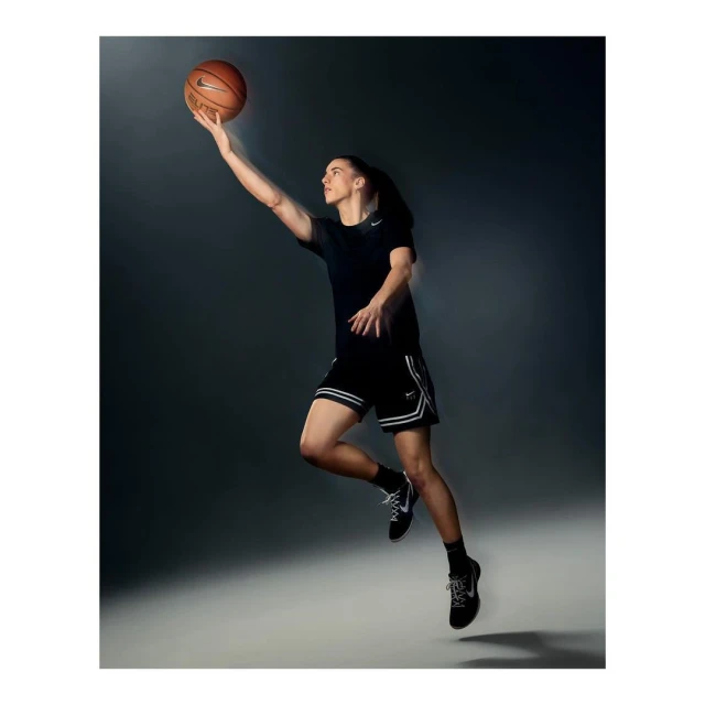 Athletic woman jumps in the air to shoot a basketball in a studio with a gray background and bright lighting. She wears Nike athletic wear.