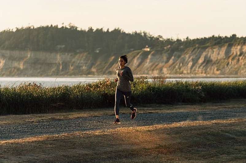 A woman runs on a path outdoors near a body of water and cliffs during the day. Fitness, active lifestyle, and nature meet in this shot.
