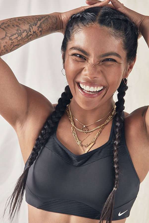 Portrait of an adult woman smiling with hands in her hair, wearing a black sports bra and gold necklaces in a light indoor setting.