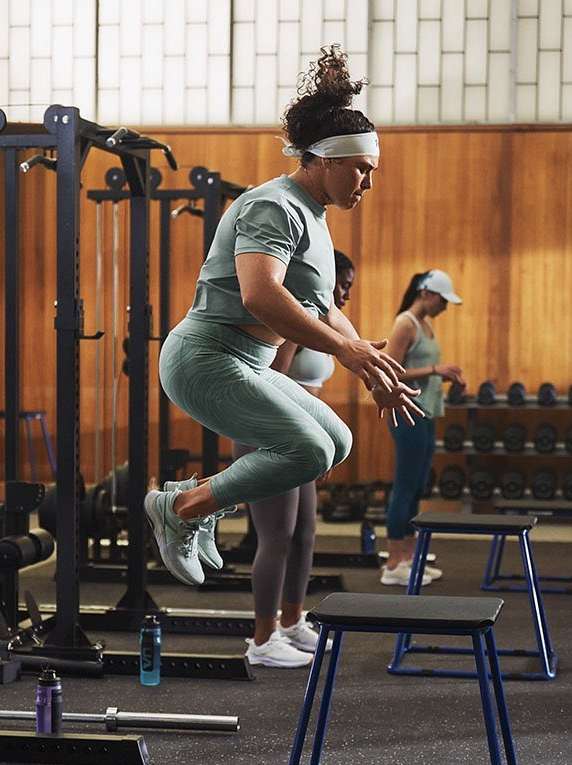 A woman jumps over a stool as part of a fitness workout, with two other people and exercise equipment in the background.
