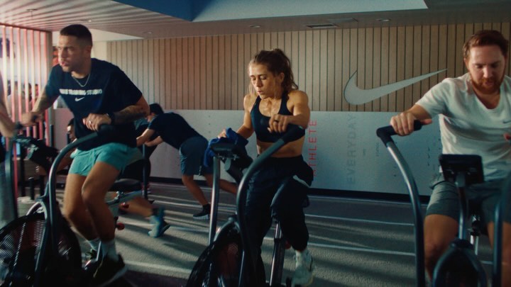 Several athletes train on stationary bikes in a modern fitness studio with natural light, wearing athletic gear and Nike branding visible.