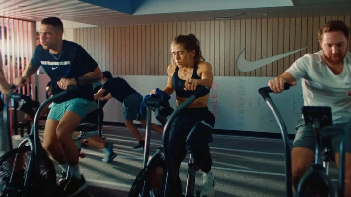 Several athletes train on stationary bikes in a modern fitness studio with natural light, wearing athletic gear and Nike branding visible.