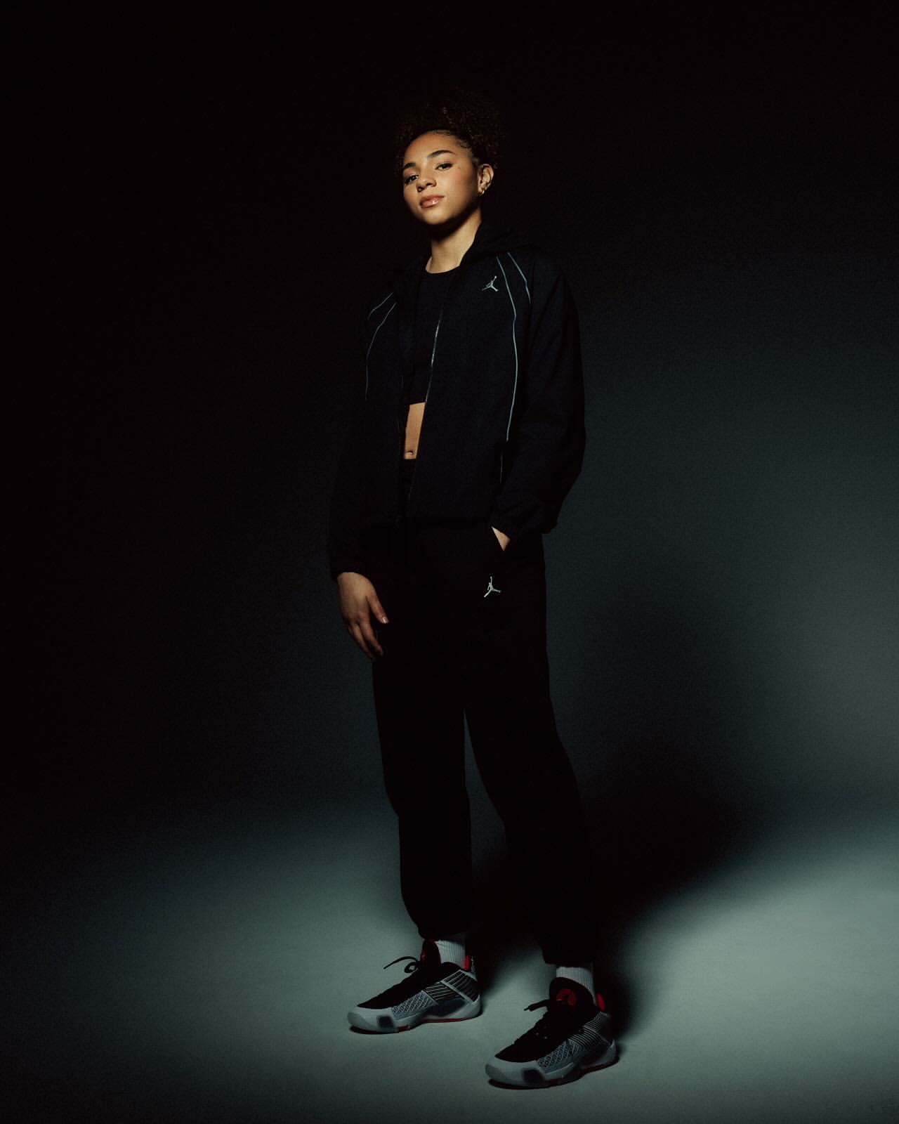 Young adult woman in a dark sports outfit with jacket and pants, posing in sneakers against a low-key, gradient gray and black background.
