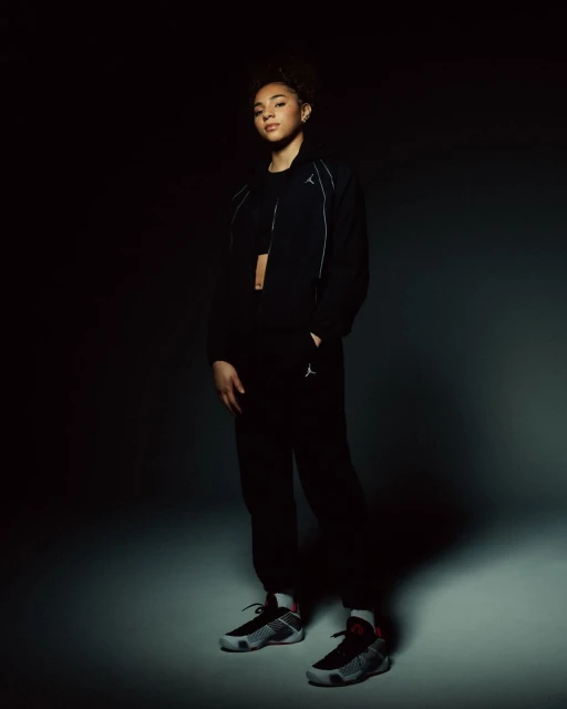 Young adult woman in a dark sports outfit with jacket and pants, posing in sneakers against a low-key, gradient gray and black background.