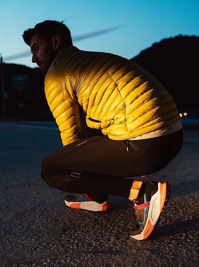 Male athlete pauses outdoors at dusk. He wears a yellow puffer jacket, black leggings, and colorful athletic shoes, with mountains visible.