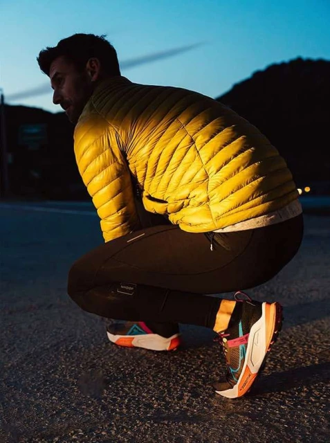 Male athlete pauses outdoors at dusk. He wears a yellow puffer jacket, black leggings, and colorful athletic shoes, with mountains visible.