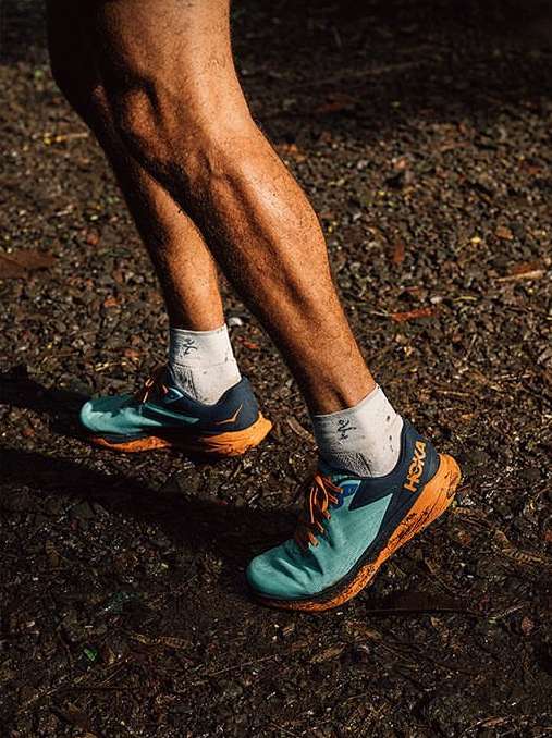 Close-up of a person's legs and feet running on a trail wearing aqua and orange Hoka running shoes and white socks with black graphics.