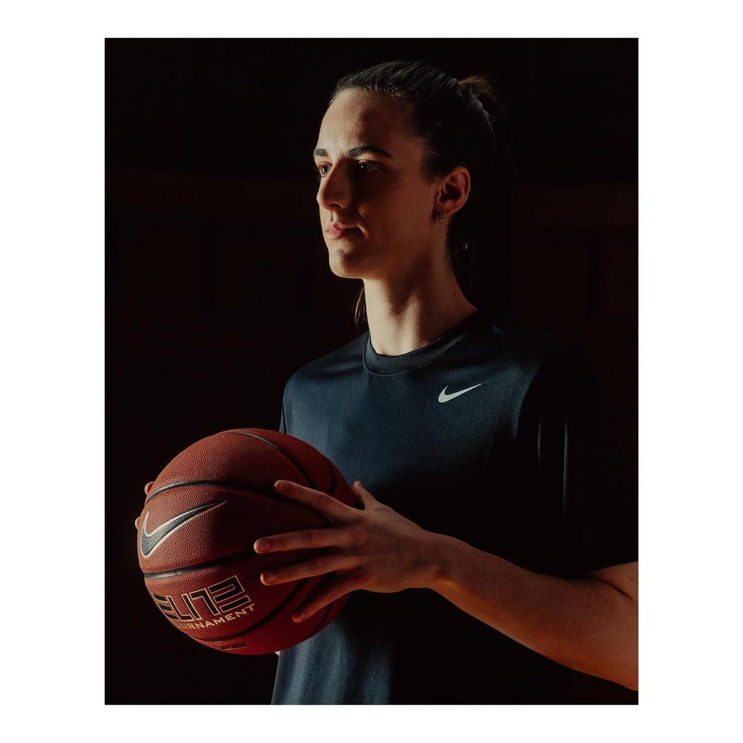Portrait of a young adult female athlete holding a basketball while wearing a navy athletic shirt with a Nike logo in a dark indoor setting.