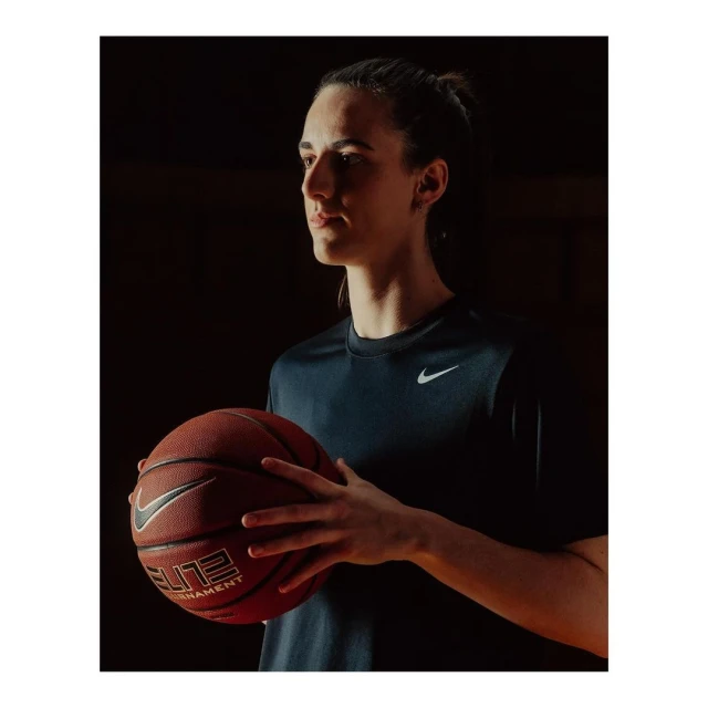 Portrait of a young adult female athlete holding a basketball while wearing a navy athletic shirt with a Nike logo in a dark indoor setting.