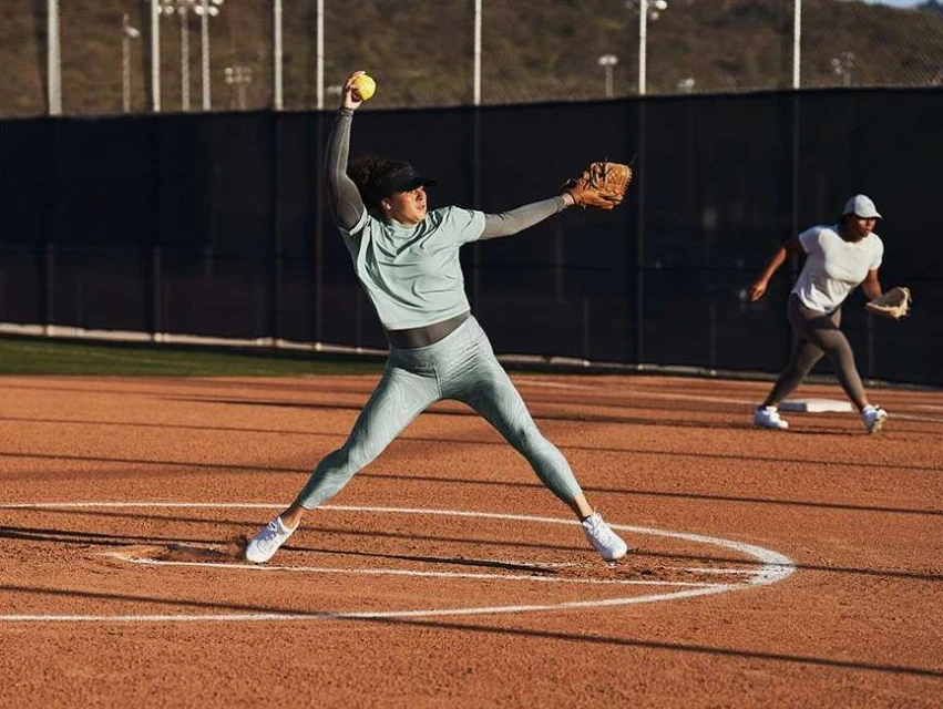 Female softball player in teal athletic wear throwing a ball on a dirt field with a second player in the background.