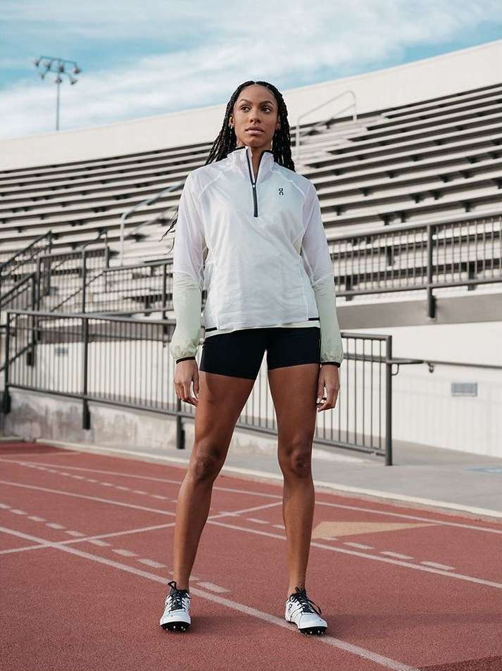 A woman in athletic wear stands confidently on a track in front of stadium seating, wearing a white jacket and running shoes.