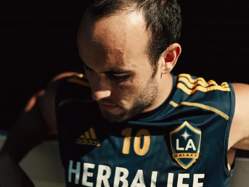 Close-up of adult male athlete in blue LA Galaxy jersey with Herbalife logo, looking down with serious expression, dark background.