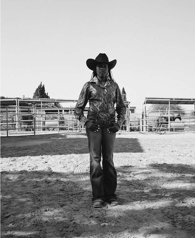 Monochrome portrait of an adult woman in cowboy hat and Western wear standing in a dirt paddock with a horse in the background.