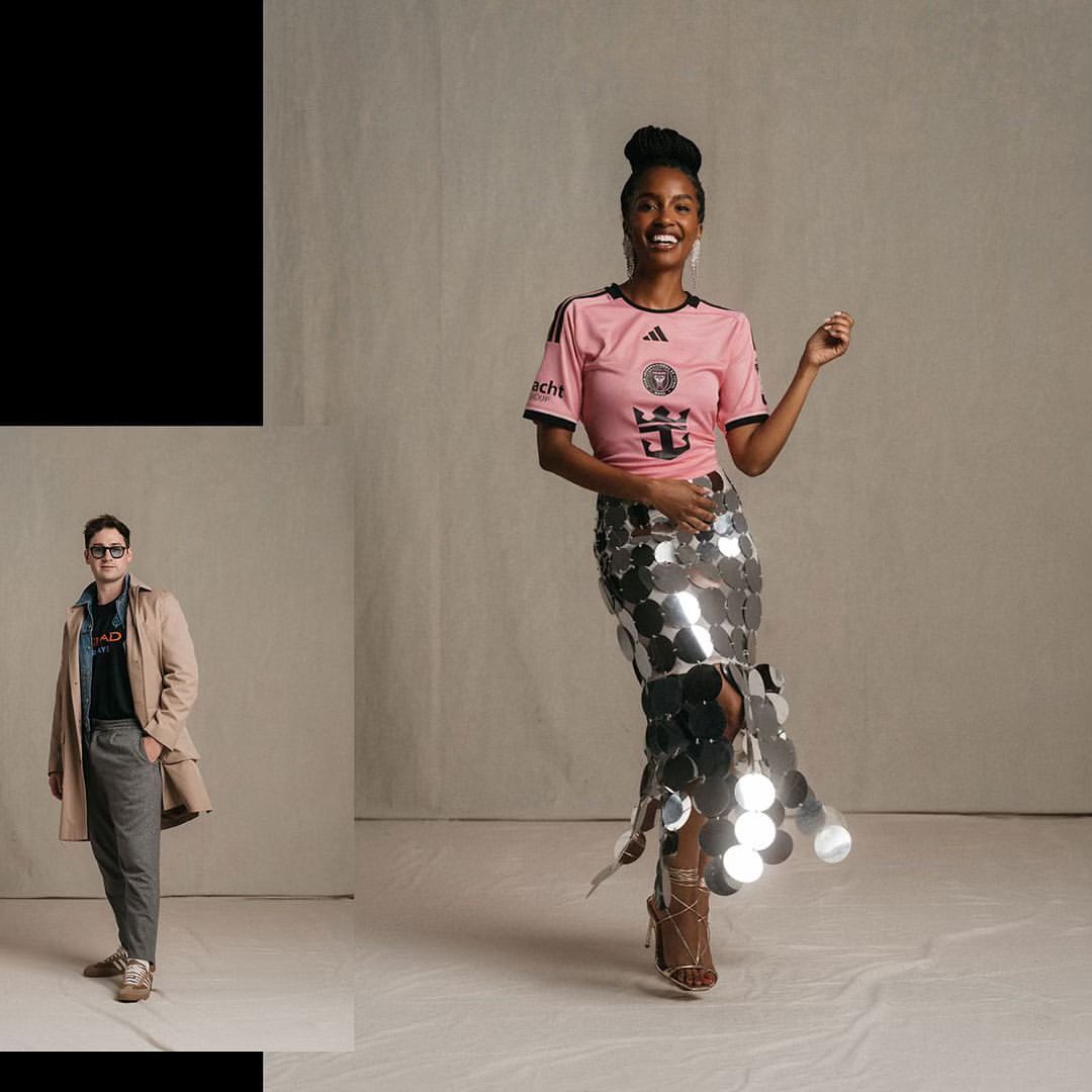 Woman in pink soccer jersey and silver skirt posing in a studio with a man in a trench coat standing to the side.