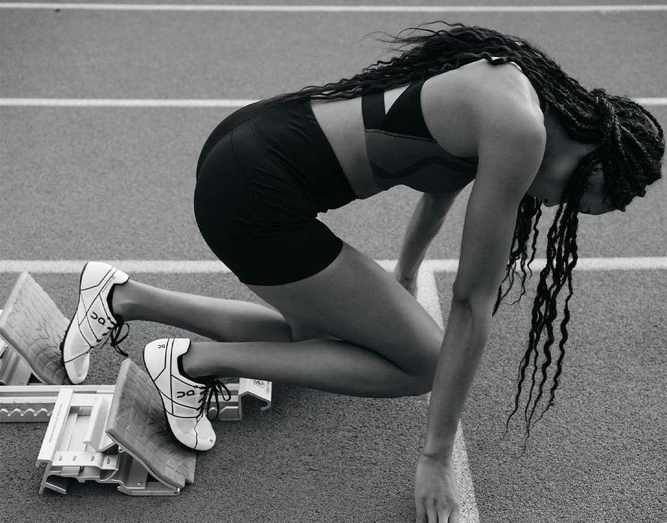 Black and white shot of a woman in athletic wear kneeling in starting blocks on a running track, preparing for a race, in a fitness setting.