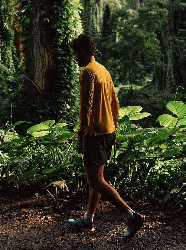 Man walking on a dirt path through a lush green forest, wearing a yellow long-sleeved shirt, black shorts, and athletic shoes.