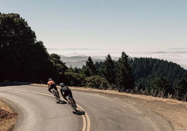 Two cyclists descending a winding asphalt road through a mountain landscape with distant fog rolling over hills.