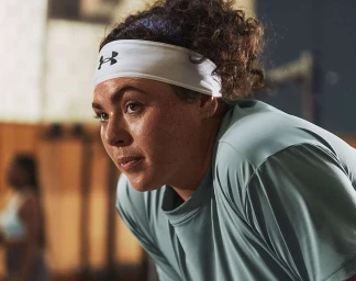 Close-up portrait of a sweating woman wearing a white Under Armour headband and sage green athletic top in a gym setting.