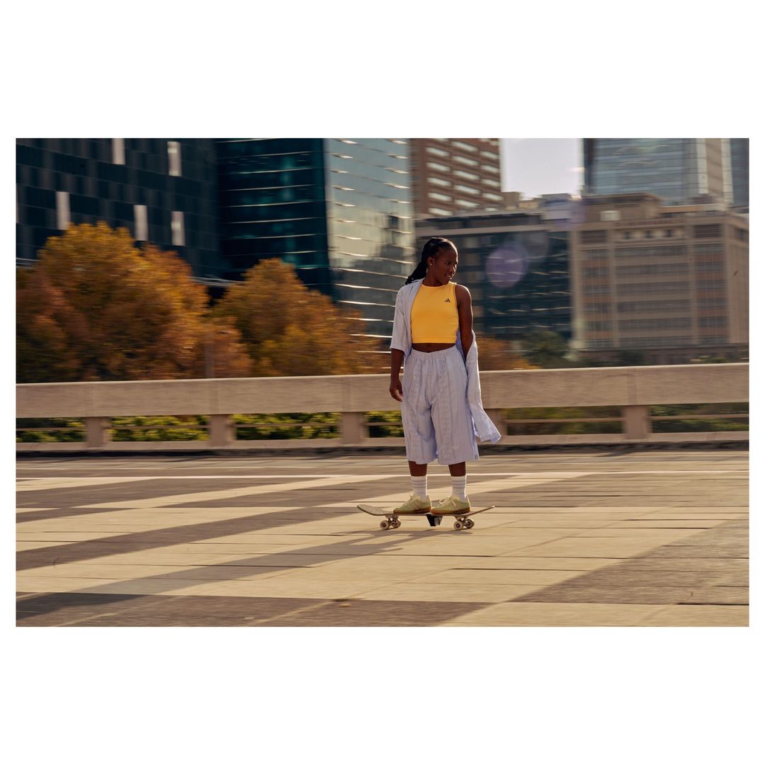 A young African-American woman is skateboarding in an urban setting. She wears a yellow top and striped shorts, with modern buildings behind.