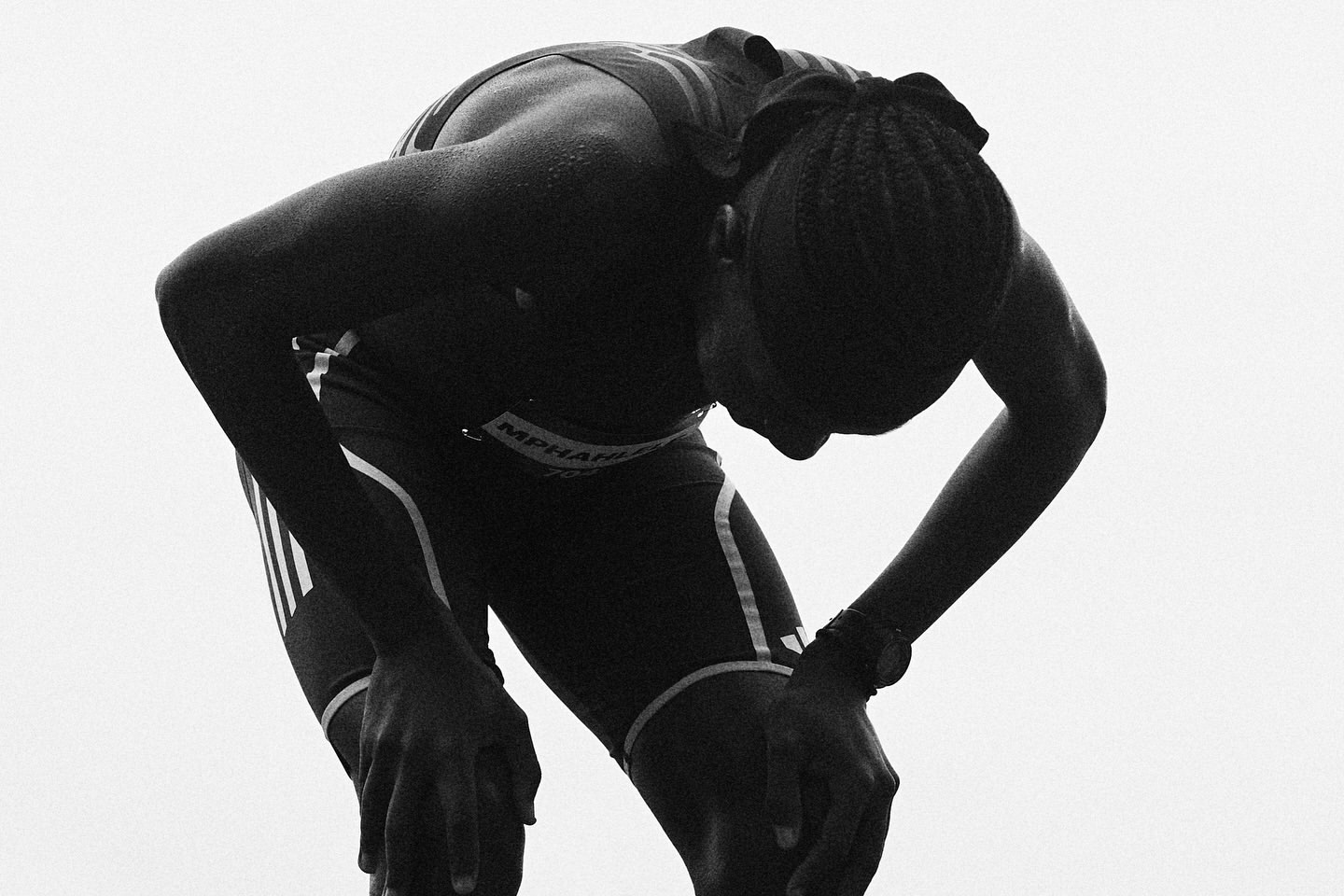 A female athlete, sweat glistening, pauses with hands on knees, head bowed, clad in sports gear, after a workout, monochrome image.