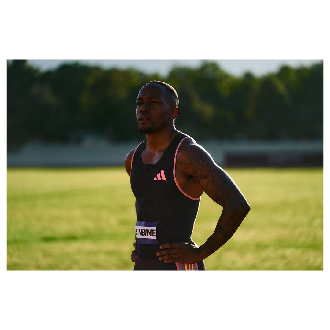 Runner pauses with hands on hips on track; African-American man in black and pink Adidas tank top, green field, and natural light.