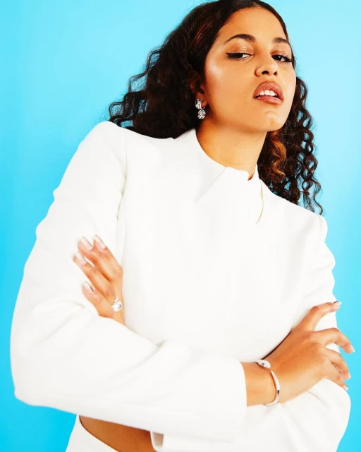 Elegant woman in white blazer posing against a light blue background in a portrait shot. Features jewelry and well-manicured nails.