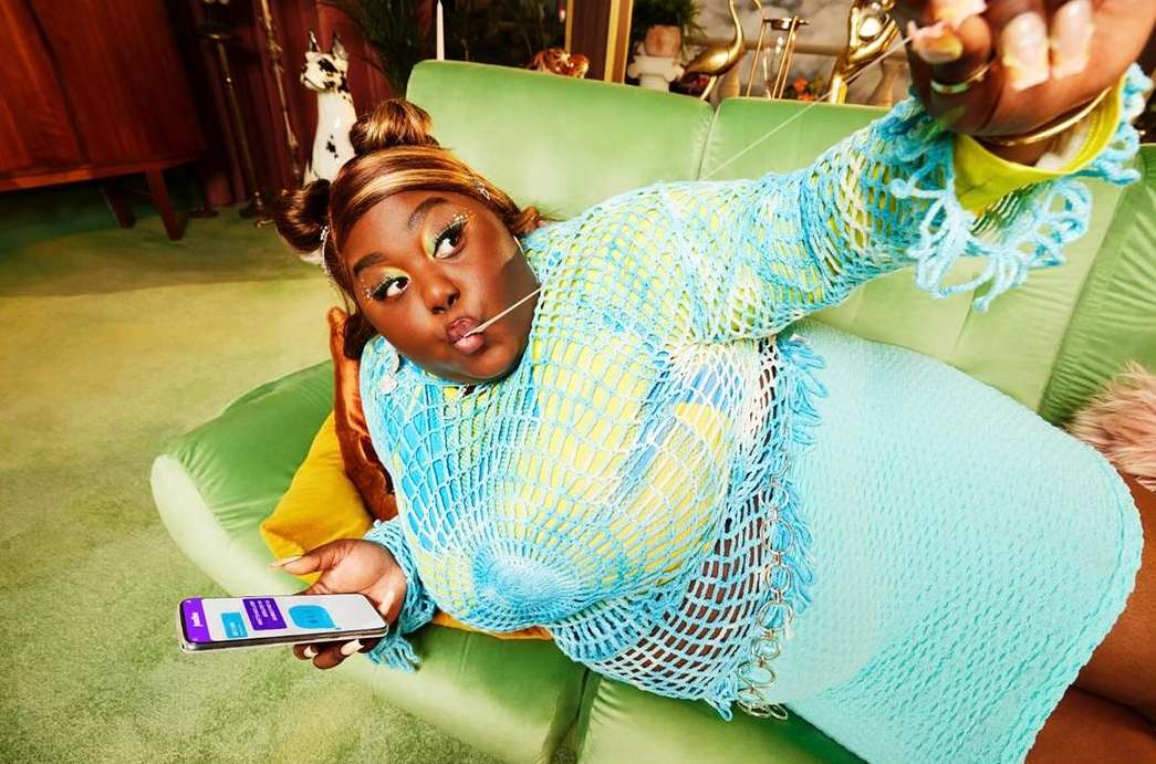 Stylish African American woman poses on a green sofa holding a smartphone wearing a blue crochet top and a matching blue skirt in a home setting.