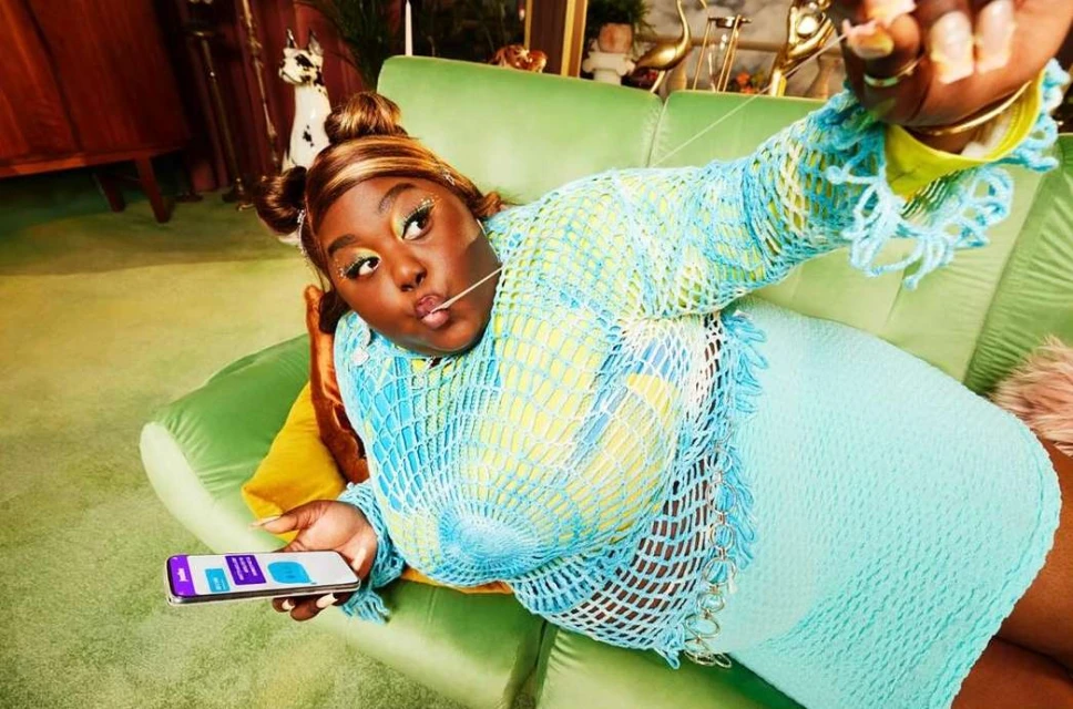 Stylish African American woman poses on a green sofa holding a smartphone wearing a blue crochet top and a matching blue skirt in a home setting.