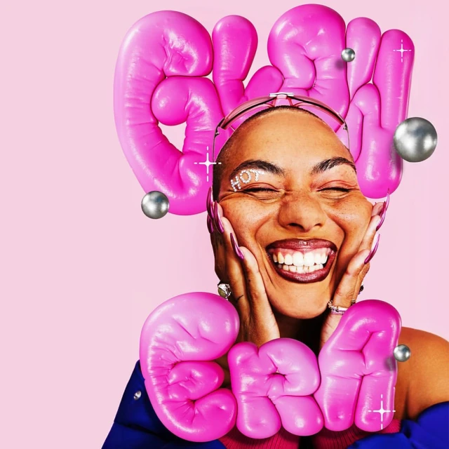 Close-up portrait of a smiling woman with pink balloon letters framing her face and silver orbs against a pastel pink backdrop.