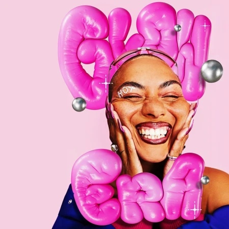 Close-up portrait of a smiling woman with pink balloon letters framing her face and silver orbs against a pastel pink backdrop.