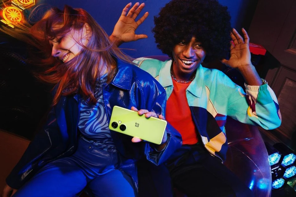 Two smiling adults, one with an afro and the other with auburn hair, are holding a light green smartphone in a colourful indoor scene.