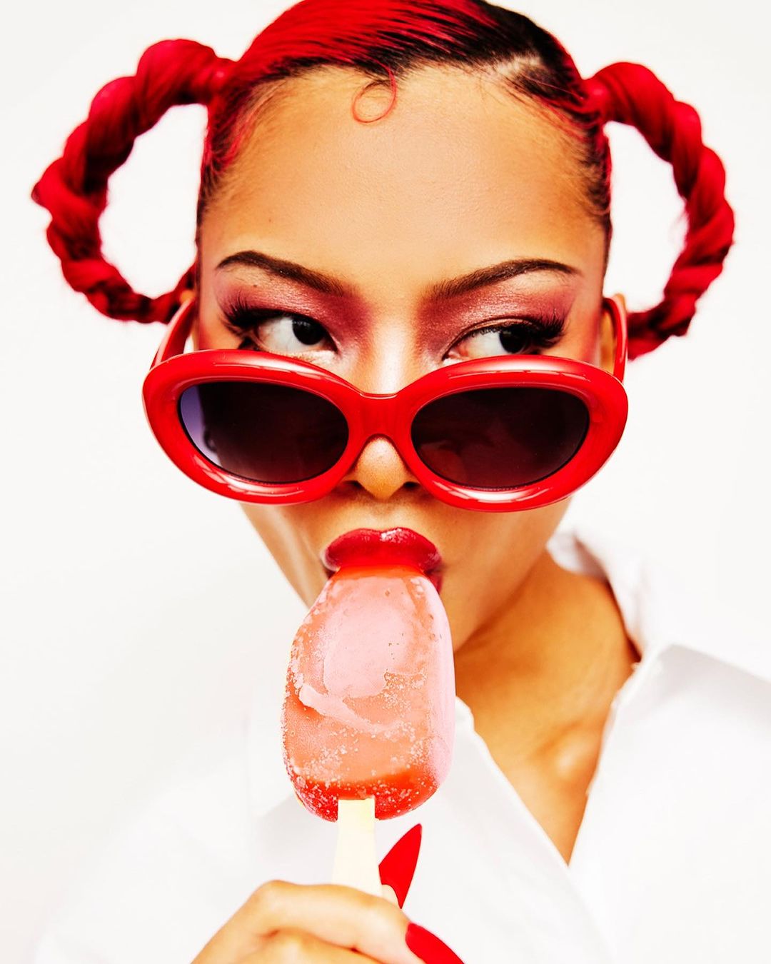 Portrait of a young woman with red hair in space buns, wearing red sunglasses and a white shirt, eating a red popsicle against a white background.