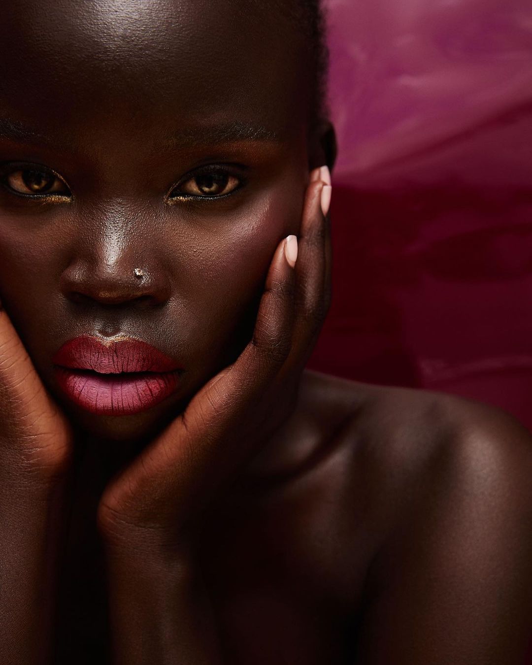 Close-up portrait of a black female model with makeup, posing with her hands framing her face against a blurred magenta background.