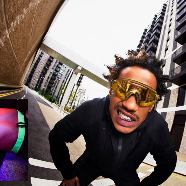 Low-angle shot of an adult man with golden sunglasses and a black jacket on a city street, with tall buildings in the background.