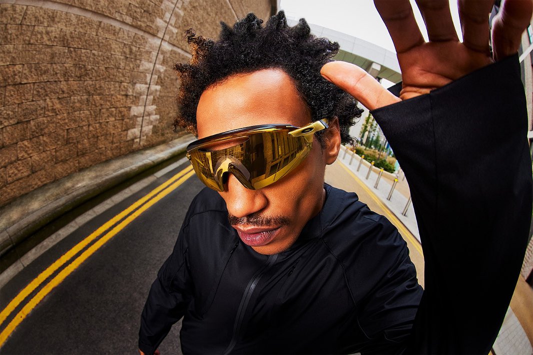 African American man in gold sunglasses and black hooded jacket gazes at the camera on a city street with an urban tunnel background.