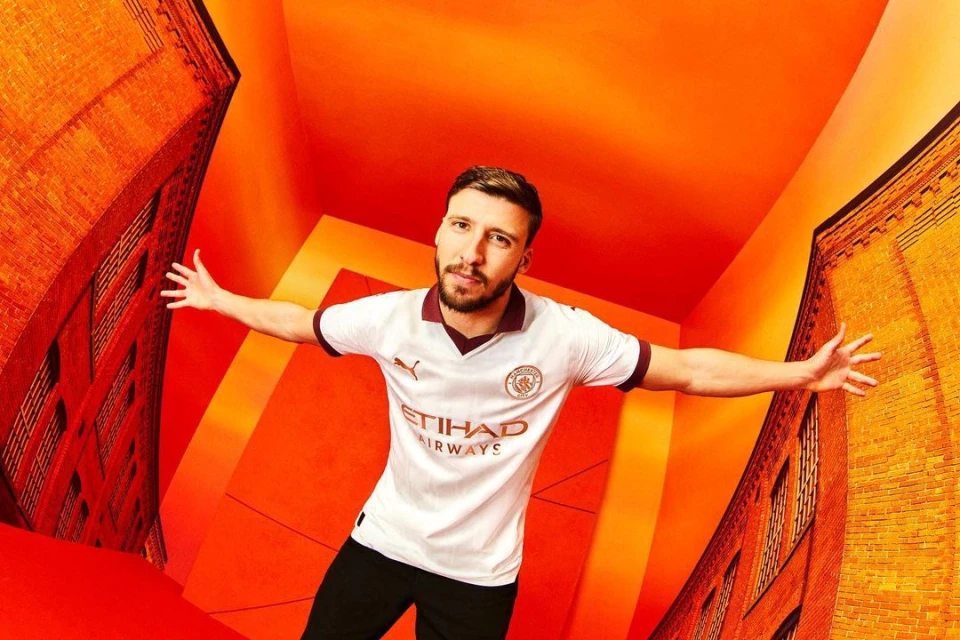 Adult male in a white soccer jersey with 'ETIHAD AIRWAYS' text, posing in front of an orange geometric background with brick building prints.