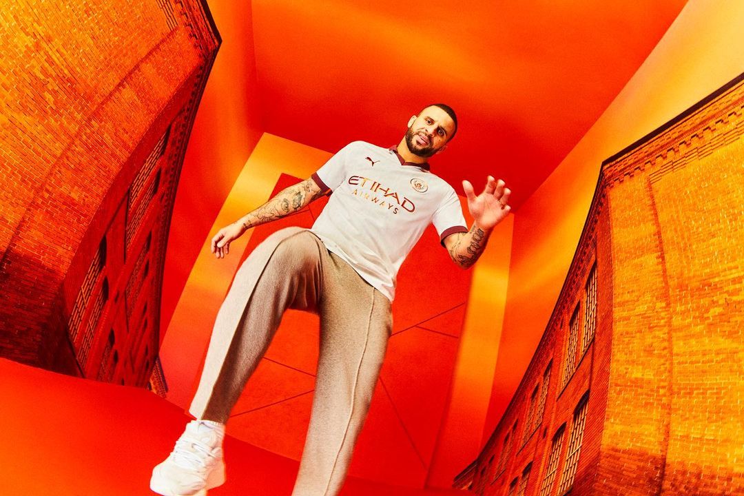 Adult male in a soccer jersey poses in a visually distorted studio with orange walls and brick-building details, captured from a low angle.