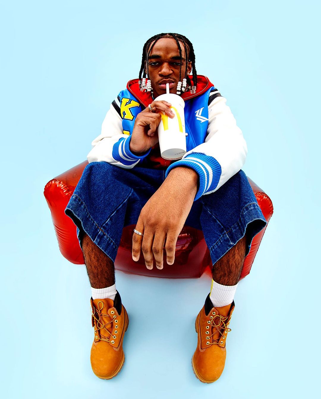 Adult man seated, drinking from a McDonald's cup, wearing a jacket, denim culottes, boots, and white socks against a solid blue background.