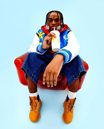 Adult man seated, drinking from a McDonald's cup, wearing a jacket, denim culottes, boots, and white socks against a solid blue background.