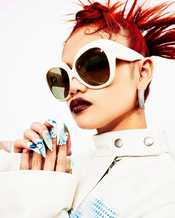 Stylish model with red hair poses in a white jacket and sunglasses, showcasing vibrant nail art against a clean white background.