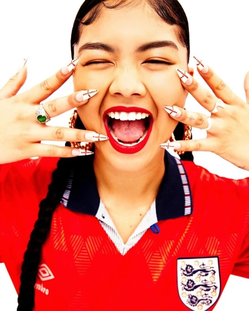 Smiling young adult woman with fair skin wearing a red soccer jersey, decorative nails, and gold earrings against a white background.