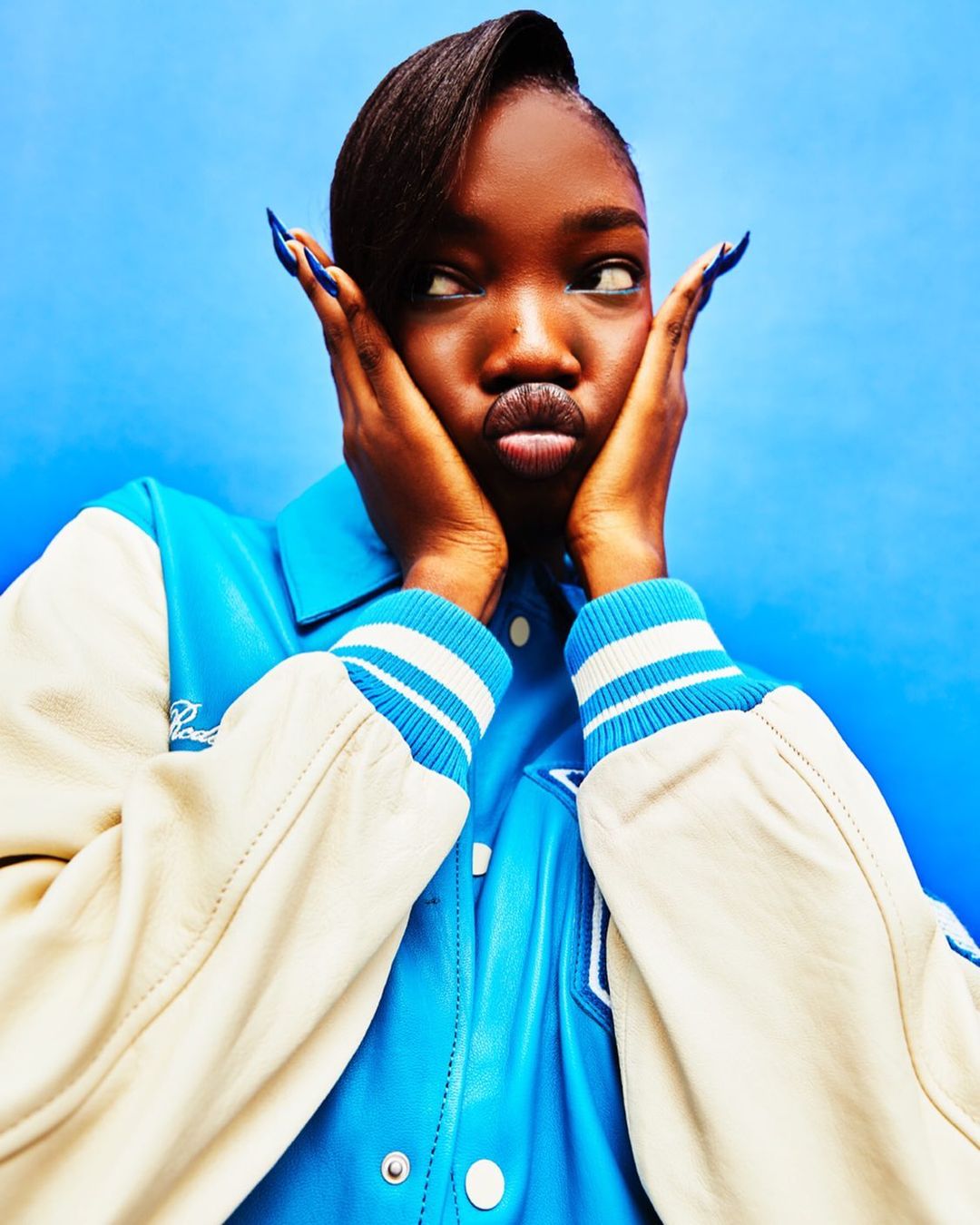 A dark-skinned female makes a funny face with her hands to her cheeks while wearing a blue-and-white varsity jacket against a blue background.