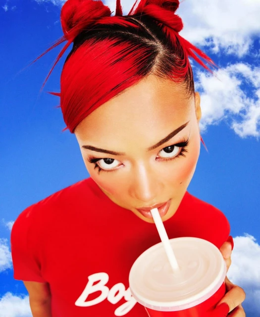 Young woman with red hair drinking from a red plastic cup through a straw against a vibrant blue sky backdrop with fluffy white clouds.