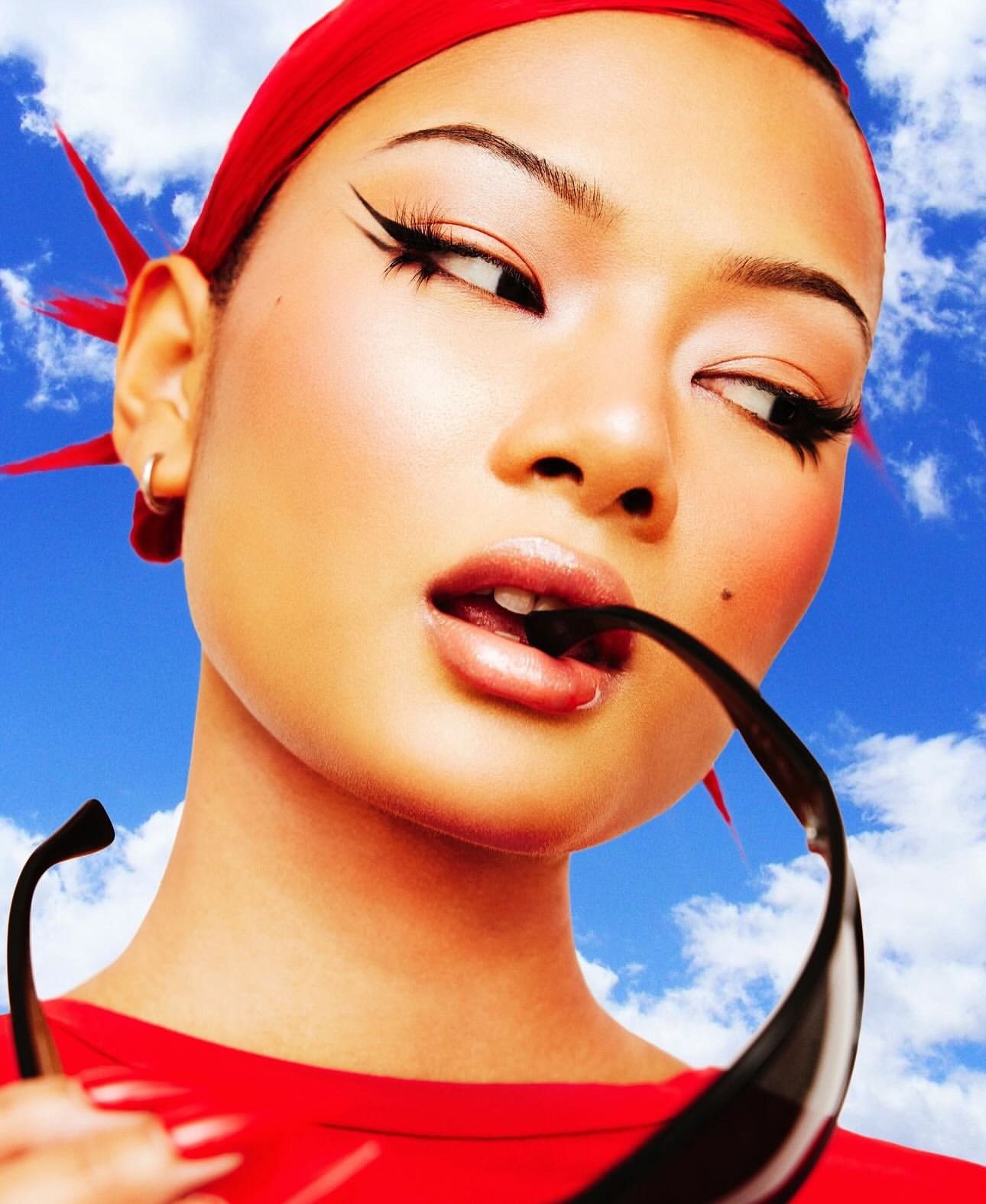 Close-up of a woman with dramatic winged eyeliner wearing a red headscarf and sunglasses with a blue sky and clouds background.