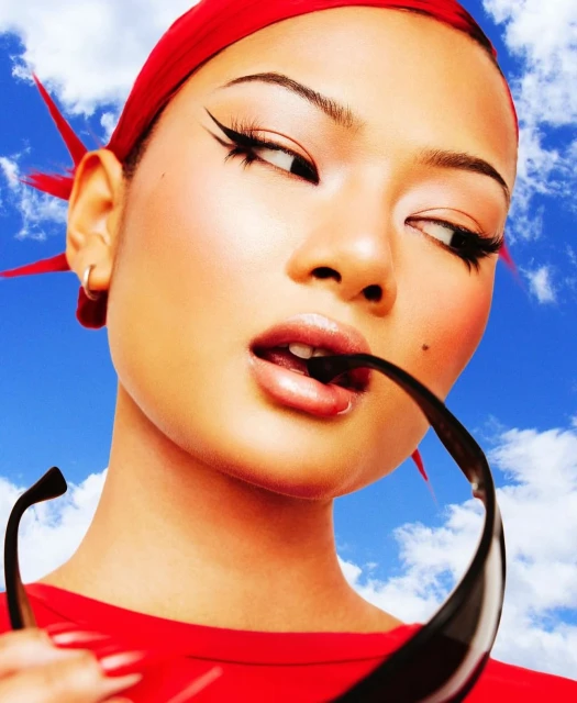 Close-up of a woman with dramatic winged eyeliner wearing a red headscarf and sunglasses with a blue sky and clouds background.