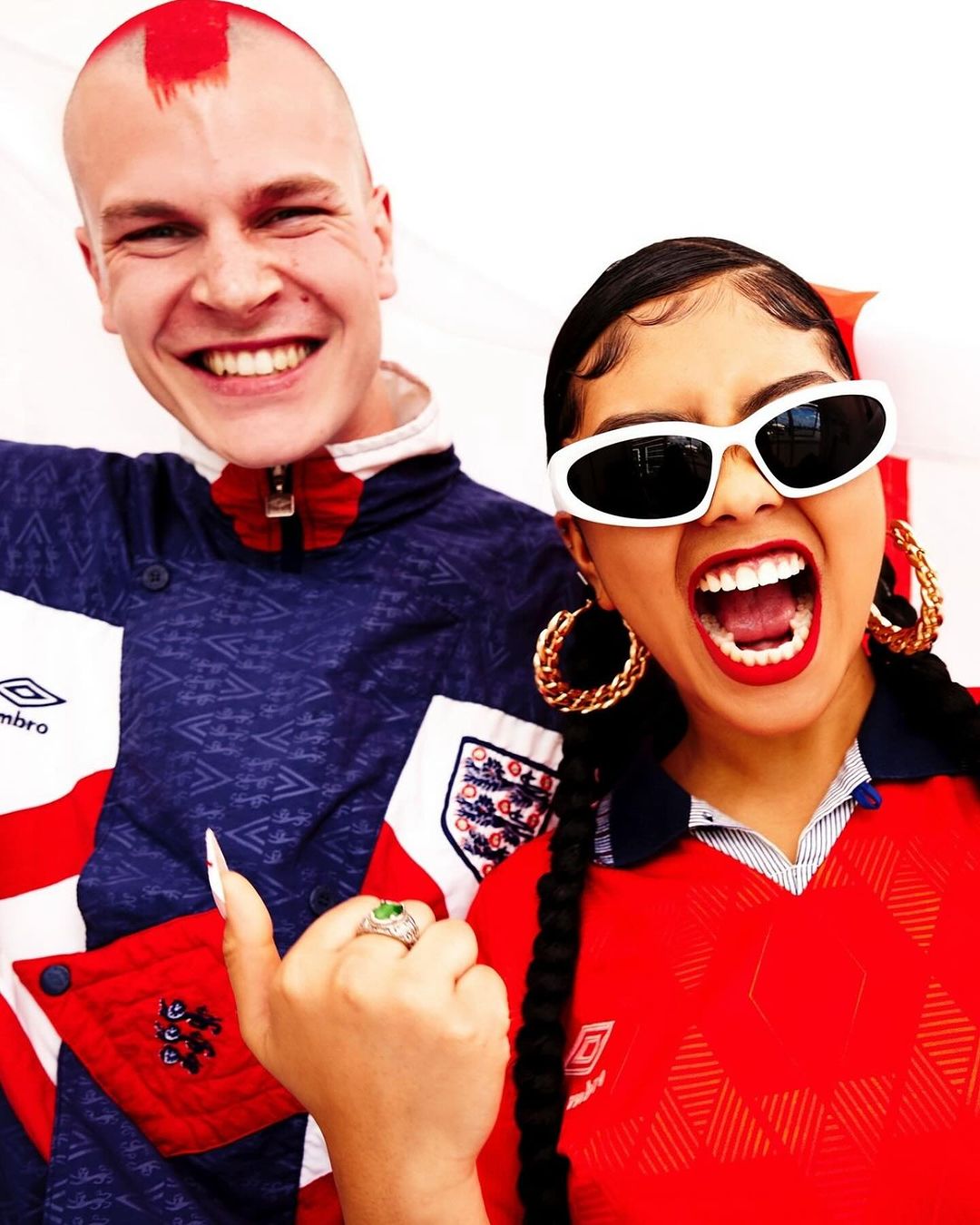 Two people wearing England soccer jerseys smile at the camera. The man has a red mohawk, and the woman is wearing white sunglasses.