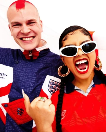 Two people wearing England soccer jerseys smile at the camera. The man has a red mohawk, and the woman is wearing white sunglasses.